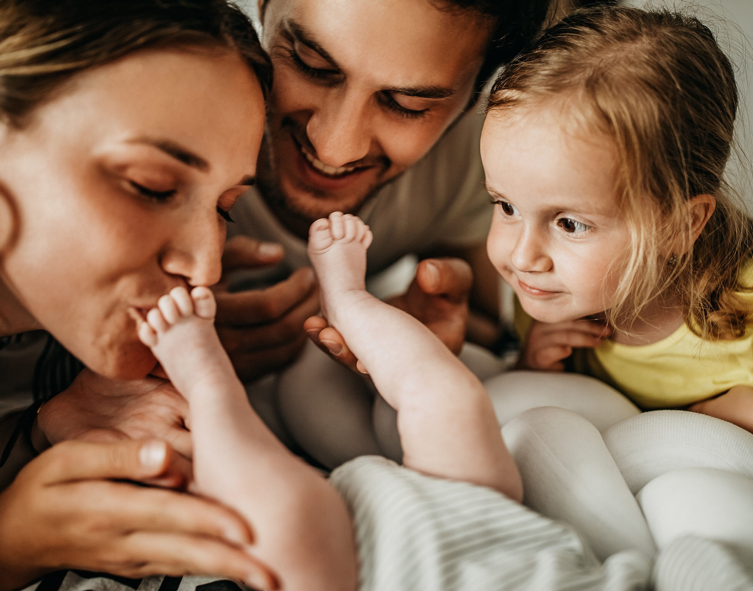 Zwei Elternteile und ein Kind im Kindergartenalter sehen sich die Füße eines auf dem Tisch liegenden Babys an.