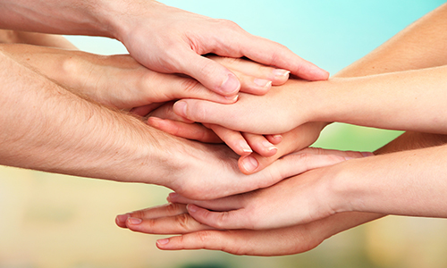 Die Arme von verschiedenen Menschen zeigen zur Mitte des Bildes, dort sind alle Hände aufeinander gelegt.