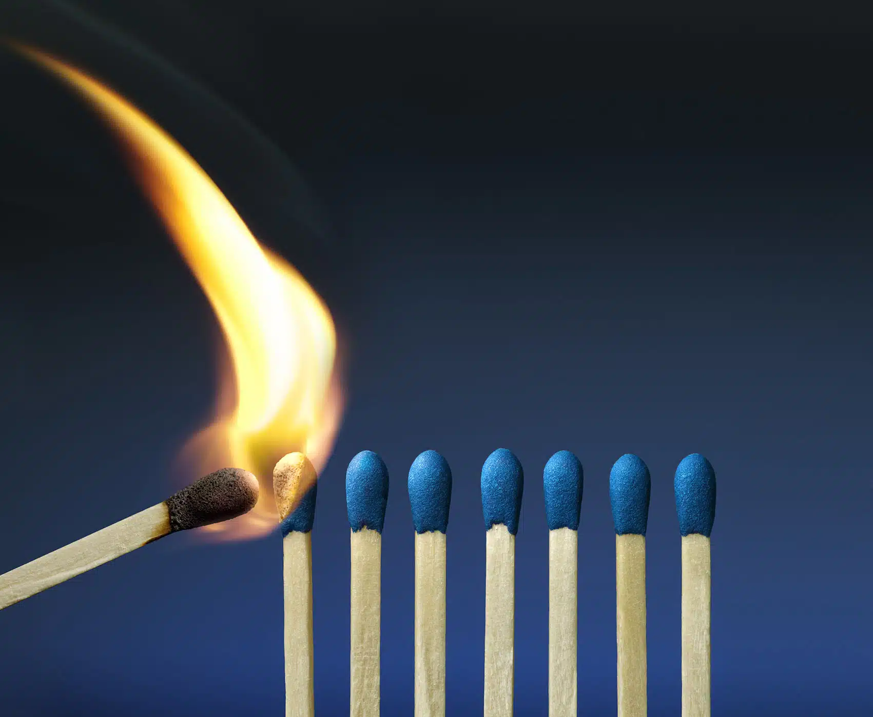 Mehrere Streichhölzer senkrecht nebeneinander aufgereiht. Das linke Streichholz wird von einem anderen Streichholz angezündet.
